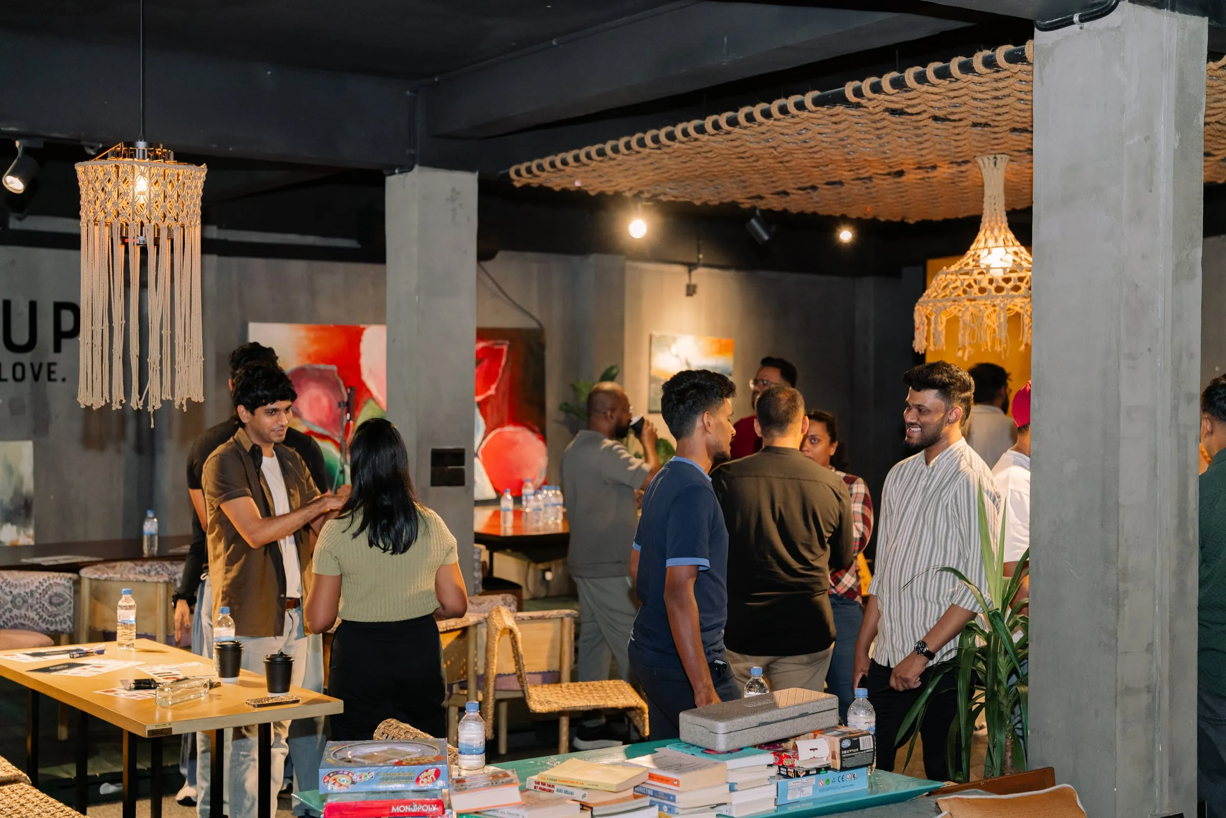 Designers standing in small groups and talking in a dimly lit café, with paintings on the concrete walls and woven lamps overhead.